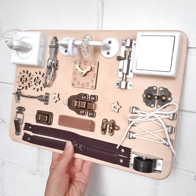 May include: A wooden activity board with various textures and activities for children. It features a clock, a zipper, a latch, a toggle, a star, a wheel, a cord, and a light switch.