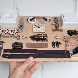 K&ouml;nnte beinhalten: Ein Holz-Besch&auml;ftigungsbrett mit verschiedenen Aktivit&auml;ten f&uuml;r Kinder, darunter ein Rei&szlig;verschluss, ein Riegel, eine Schnalle, eine Uhr, ein Griff, ein Knebelverschluss, ein Klettverschluss und ein Zahnradpaar. Das Brett ist aus hellem Holz und hat eine nat&uuml;rliche Oberfl&auml;che.