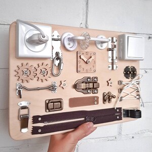 May include: A wooden activity board with various textures and activities for children. It features a clock, a zipper, a latch, a toggle switch, a door, a knob, and gears. The board is painted in a light wood color.