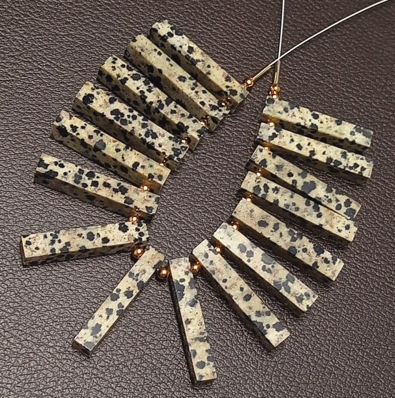 May include: A strand of dalmatian jasper beads, each bead is a rectangular prism with a black and white speckled pattern. The beads are strung on a wire with gold-colored spacers between each bead.