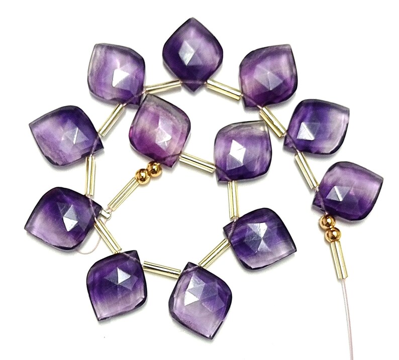 May include: A strand of purple amethyst gemstone beads with gold spacers. The beads are faceted and have a square shape with rounded corners.