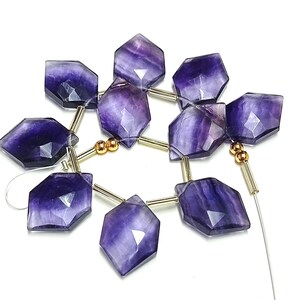 Puede incluir: Un hilo de cuentas facetadas hexagonales de fluorita en tonos morado intenso y lavanda. Las cuentas est&aacute;n ensartadas con elementos met&aacute;licos dorados y un hilo fino y transparente, creando una disposici&oacute;n floral. Las cuentas est&aacute;n pulidas y tienen un acabado brillante.