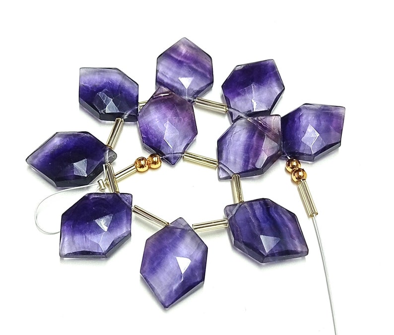 Puede incluir: Un hilo de cuentas facetadas hexagonales de fluorita en tonos morado intenso y lavanda. Las cuentas est&aacute;n ensartadas con elementos met&aacute;licos dorados y un hilo fino y transparente, creando una disposici&oacute;n floral. Las cuentas est&aacute;n pulidas y tienen un acabado brillante.