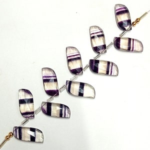 May include: A strand of purple and white fluorite gemstone beads. The beads are faceted and have a rectangular shape. The beads are strung on a silver wire with small silver spacers between each bead.