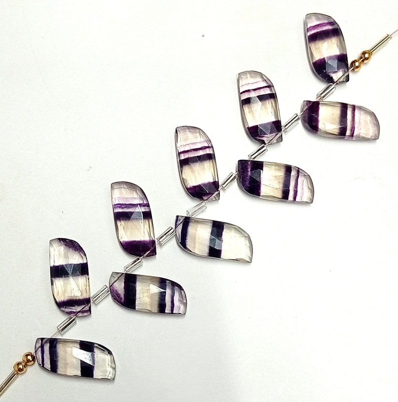 May include: A strand of purple and white fluorite gemstone beads. The beads are faceted and have a rectangular shape. The beads are strung on a silver wire with small silver spacers between each bead.