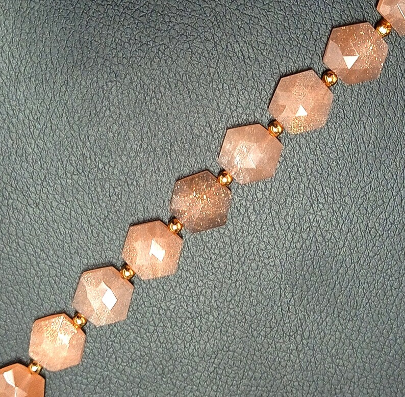 May include: A strand of faceted sunstone beads with a gold-toned metal spacer between each bead. The beads are a light peach color with a shimmery, iridescent finish.