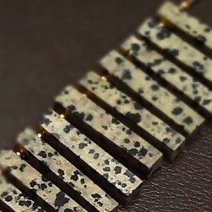 May include: A gold necklace with a series of rectangular dalmatian jasper stones. The stones are a light beige color with black spots.