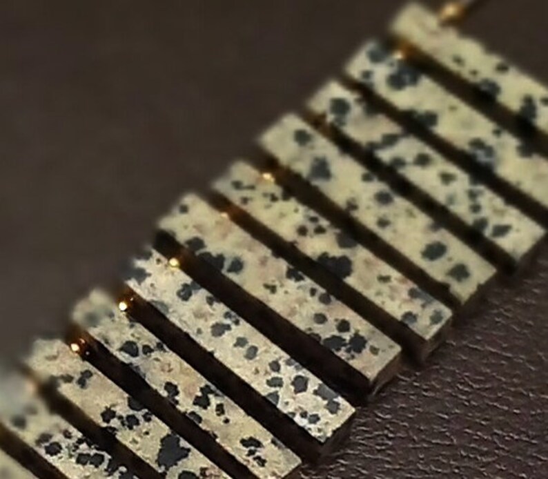 May include: A gold necklace with a series of rectangular dalmatian jasper stones. The stones are a light beige color with black spots.