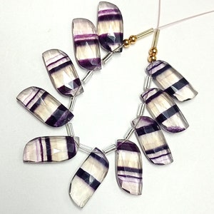 May include: A strand of faceted fluorite beads with purple and white stripes. The beads are strung on a clear thread with silver and gold spacers.