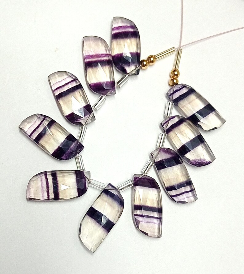 May include: A strand of faceted fluorite beads with purple and white stripes. The beads are strung on a clear thread with silver and gold spacers.
