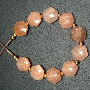 May include: A strand of sunstone beads with a hexagonal shape. The beads are a light orange color with gold flecks. The beads are strung on a thin gold wire with small gold beads between each sunstone bead.