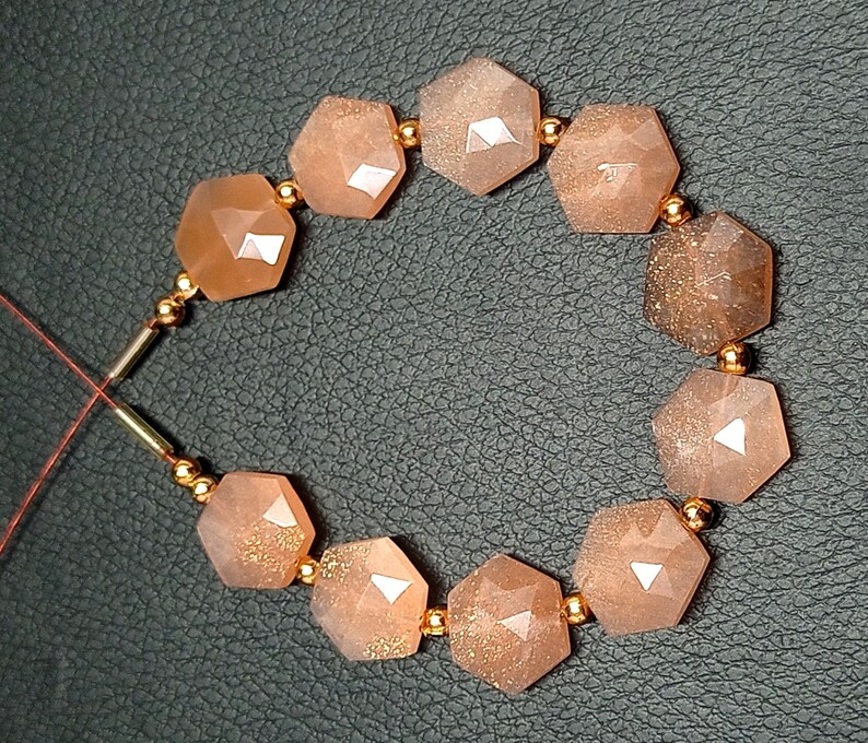 May include: A strand of sunstone beads with a hexagonal shape. The beads are a light orange color with gold flecks. The beads are strung on a thin gold wire with small gold beads between each sunstone bead.