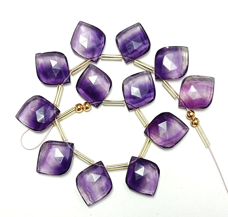 May include: A strand of purple faceted gemstone beads with gold spacers. The beads are strung on a white thread.