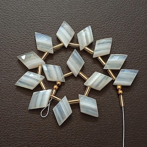 May include: A collection of diamond-shaped, light gray and white gemstone beads strung together with gold-colored metal bars and small gold beads. The gemstones have a striped pattern. The beads are arranged in a circular pattern.