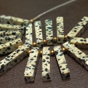 May include: A close-up of a necklace made with white and black speckled stone beads and gold spacers. The beads are arranged in a fan shape.