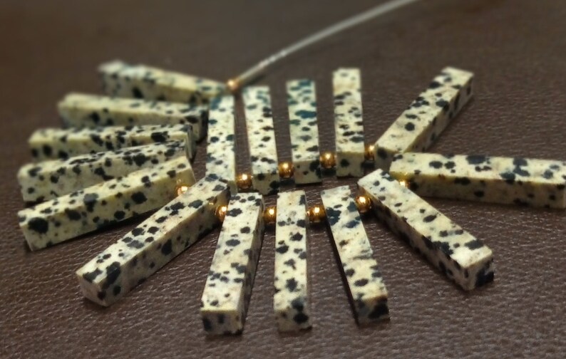 May include: A close-up of a necklace made with white and black speckled stone beads and gold spacers. The beads are arranged in a fan shape.