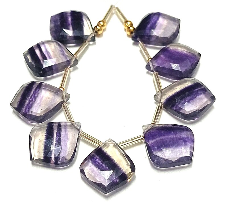 May include: A strand of purple and white fluorite gemstone beads, each bead is faceted and has a unique pattern. The beads are strung on a gold wire and are ready to be made into jewelry.