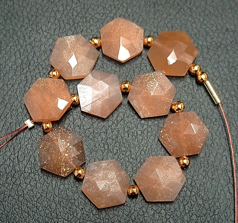 May include: A strand of 10 faceted, hexagonal sunstone beads with a copper-toned metallic sheen. The beads are strung on a copper-toned wire with small copper-toned beads between each sunstone bead.
