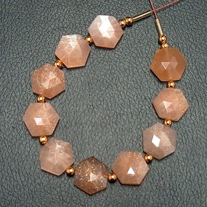 May include: A strand of sunstone beads with a faceted, hexagonal shape. The beads are a light peach color with a shimmery, glittery finish. The beads are strung on a thin, gold-colored wire.