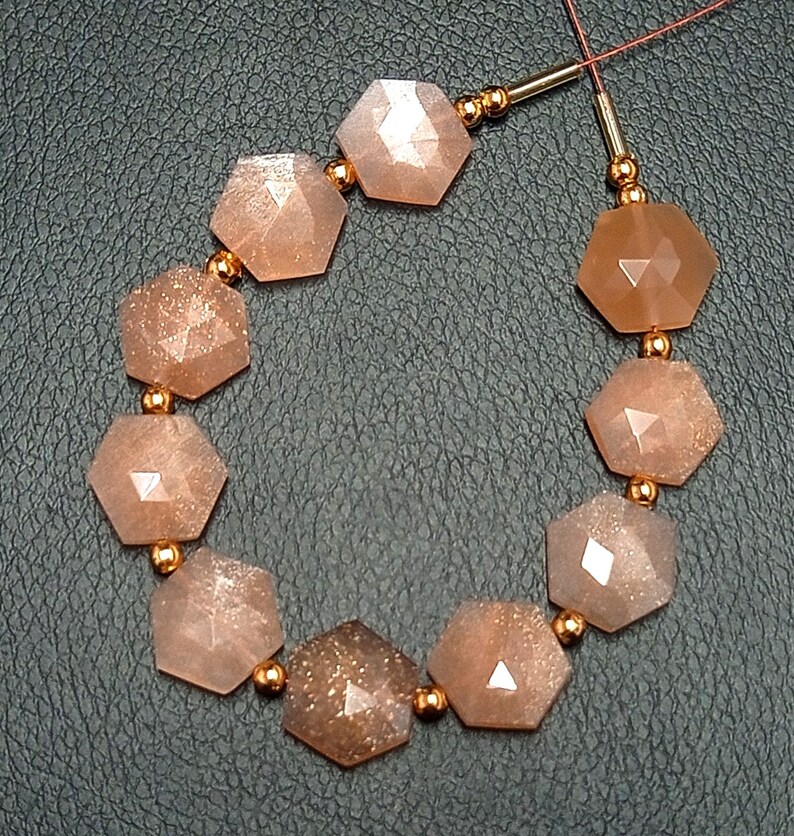 May include: A strand of sunstone beads with a faceted, hexagonal shape. The beads are a light peach color with a shimmery, glittery finish. The beads are strung on a thin, gold-colored wire.