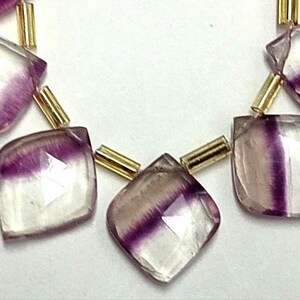 Op de afbeelding: Een streng facetgeslepen fluorite edelsteen kralen in paars en wit, gescheiden door gouden spacers. De kralen zijn vierkant gesneden met afgeronde hoeken.