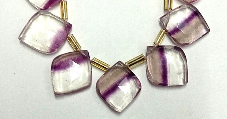 Op de afbeelding: Een streng facetgeslepen fluorite edelsteen kralen in paars en wit, gescheiden door gouden spacers. De kralen zijn vierkant gesneden met afgeronde hoeken.
