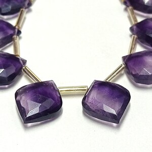 May include: A strand of purple amethyst gemstone beads, each bead is faceted and has a diamond shape. The beads are strung on a gold wire.