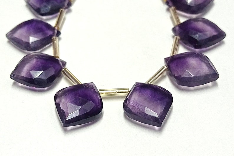 May include: A strand of purple amethyst gemstone beads, each bead is faceted and has a diamond shape. The beads are strung on a gold wire.