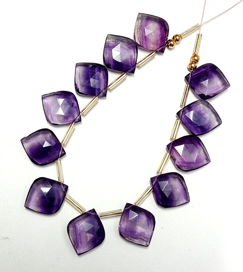 May include: A strand of purple amethyst gemstone beads with faceted, square-shaped beads. The beads are strung on gold-colored wire with small gold spacers between each bead.