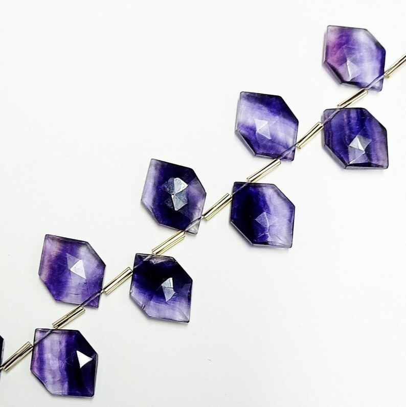 Puede incluir: Cuentas de piedras preciosas de fluorita p&uacute;rpura y blanca facetadas ensartadas. Las cuentas en forma de diamante tienen un acabado pulido y est&aacute;n ensartadas en un alambre fino de color plateado. El fondo es blanco liso.