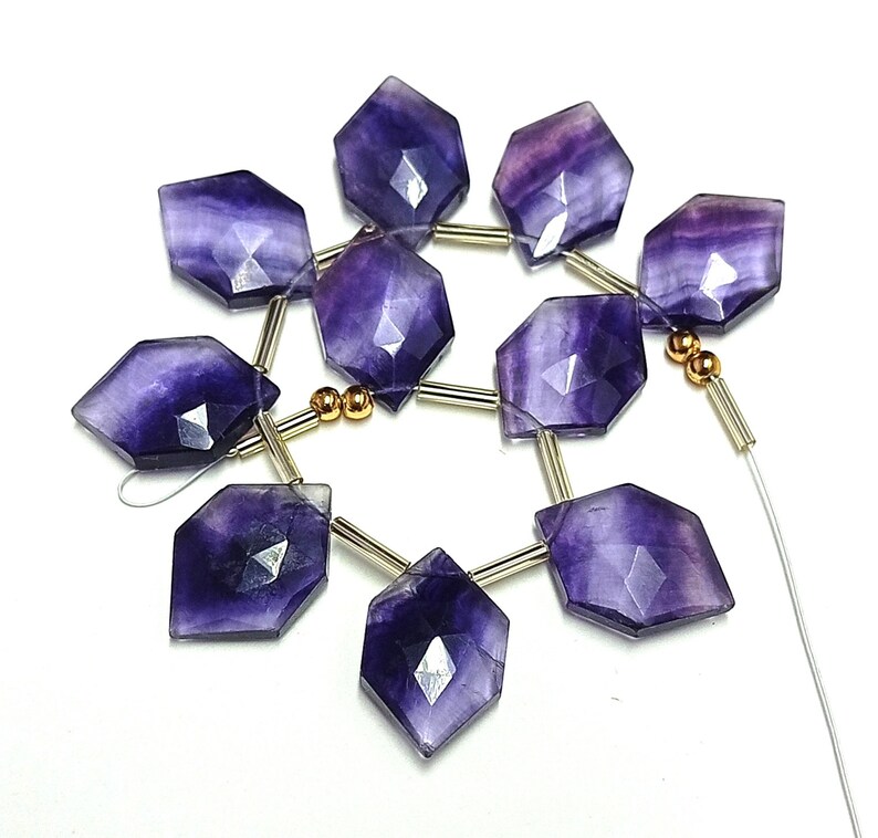 Puede incluir: Un collar de cuentas de fluorita p&uacute;rpura facetadas y hexagonales. Cada cuenta es de color violeta intenso con vetas moradas y blancas m&aacute;s claras. Las cuentas est&aacute;n ensartadas en un alambre plateado con detalles dorados.