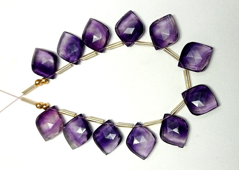 May include: A strand of purple amethyst gemstone beads with a faceted, teardrop shape. The beads are strung on a gold wire with gold spacers.