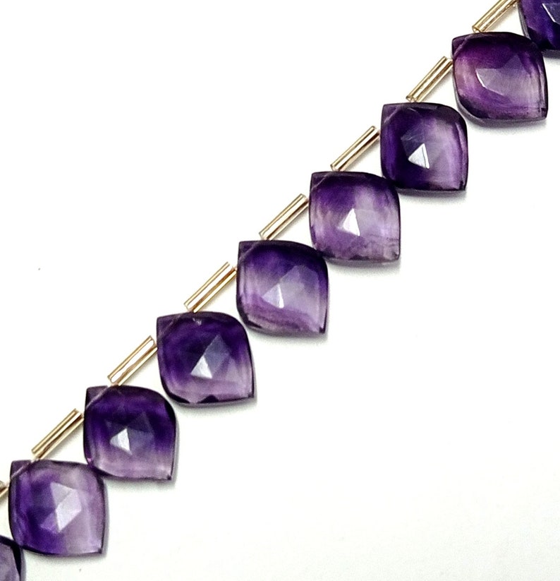 May include: A strand of faceted amethyst gemstone beads, each bead is a teardrop shape with a pointed tip and a flat base. The beads are strung on a gold wire.