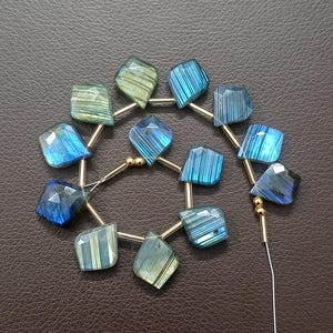 May include: A strand of faceted labradorite gemstone beads in shades of blue and green. The beads are strung on gold-colored wire, with a dark brown background. The gemstones have a geometric shape and are ready for jewelry making.