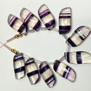 May include: A strand of purple and white striped fluorite gemstone beads. The beads are faceted and have a smooth, polished finish. The beads are strung on a clear elastic cord.