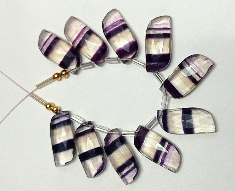 May include: A strand of purple and white striped fluorite gemstone beads. The beads are faceted and have a smooth, polished finish. The beads are strung on a clear elastic cord.