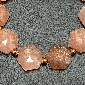 May include: A bracelet made of light brown sunstone beads with a hexagonal shape. The beads are connected with small gold-colored metal beads.