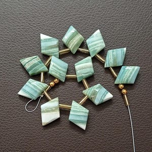 May include: A strand of gemstone beads featuring a diamond shape with a mix of green, white, and blue hues. The beads are strung together with gold-colored metal bars and small gold beads. The gemstones have a striped pattern.