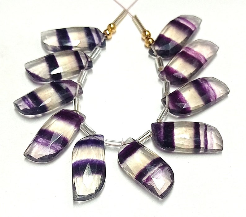 May include: A strand of purple and white striped fluorite gemstone beads. The beads are faceted and have a smooth, polished finish. The beads are strung on a silver wire with gold spacers.