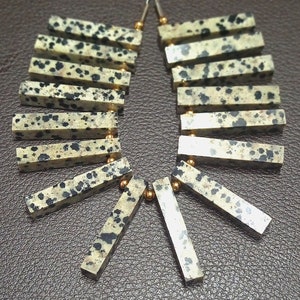May include: A strand of 18 dalmatian jasper beads, each bead is a rectangular prism with a light brown base and black spots. The beads are strung on a gold wire with gold spacers between each bead.