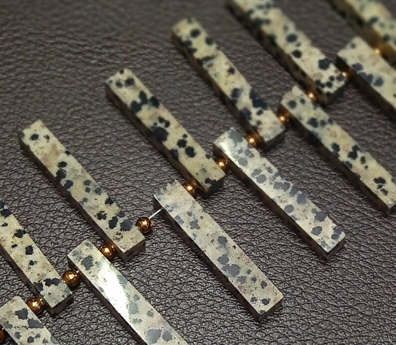 May include: A close-up of a string of brown and black speckled stone beads separated by gold spacers. The beads are rectangular in shape and have a natural, rough texture.