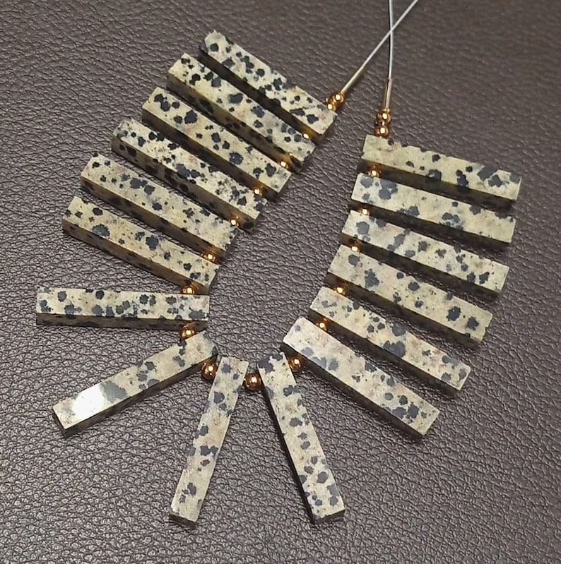 May include: A strand of dalmatian jasper beads, each bead is a rectangular shape with a light brown base and black spots. The beads are strung on a silver wire with gold spacers between each bead.