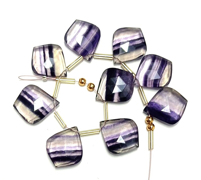 May include: A strand of purple and white fluorite gemstone beads with gold spacers. The beads are faceted and have a teardrop shape.