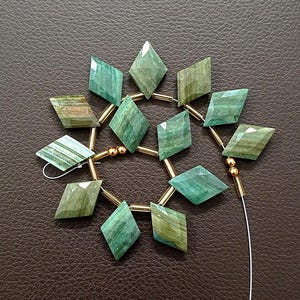 May include: A strand of green and gold gemstone beads. The beads are diamond-shaped with a faceted surface, strung together with gold-colored metal and a white string. The gemstones have varying shades of green.