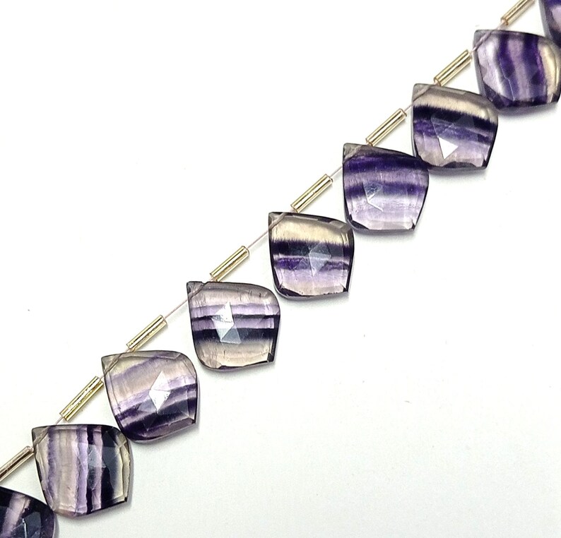 May include: A strand of purple and white striped fluorite gemstone beads. The beads are faceted and have a teardrop shape. The beads are strung on a silver wire with gold spacers.