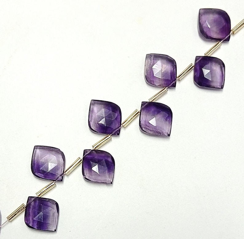 May include: A strand of amethyst gemstone beads, each bead is faceted and has a square shape. The beads are strung on a gold wire.