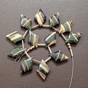 May include: Close-up of a strand of gemstone beads. The beads are diamond-shaped, with a mix of green, tan, and cream colors. They are strung together with gold-colored metal and a thin white string.