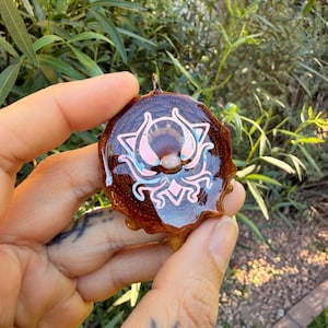 May include: A handcrafted pendant with a unique design. The pendant features a dark brown, natural wood base with a glossy, clear resin coating. A light pink and blue design is centered on the pendant. The pendant is held by a person.