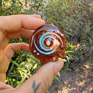 May include: A brown resin pendant with a spiral design in blue, pink, and white. The pendant has a small, round, light-colored bead in the center and is held up by a small gold-colored loop.
