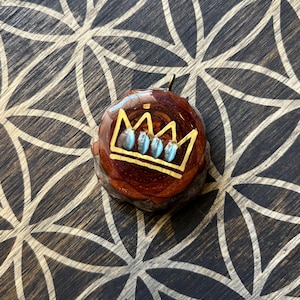 May include: A round wooden pendant with a gold crown design and blue gemstones. The pendant is on a dark wood background.